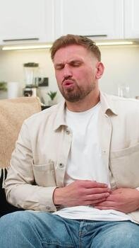 Young man at home presses hands to stomach feels cramps and pain after heavy fatty food and breathes. Caucasian guy on sofa curls slightly and shows nausea spasm and low mood while waiting for relief video